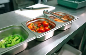 Stainless steel containers holding fresh tomatoes, sliced vegetables and kitchen utensils on a countertop.