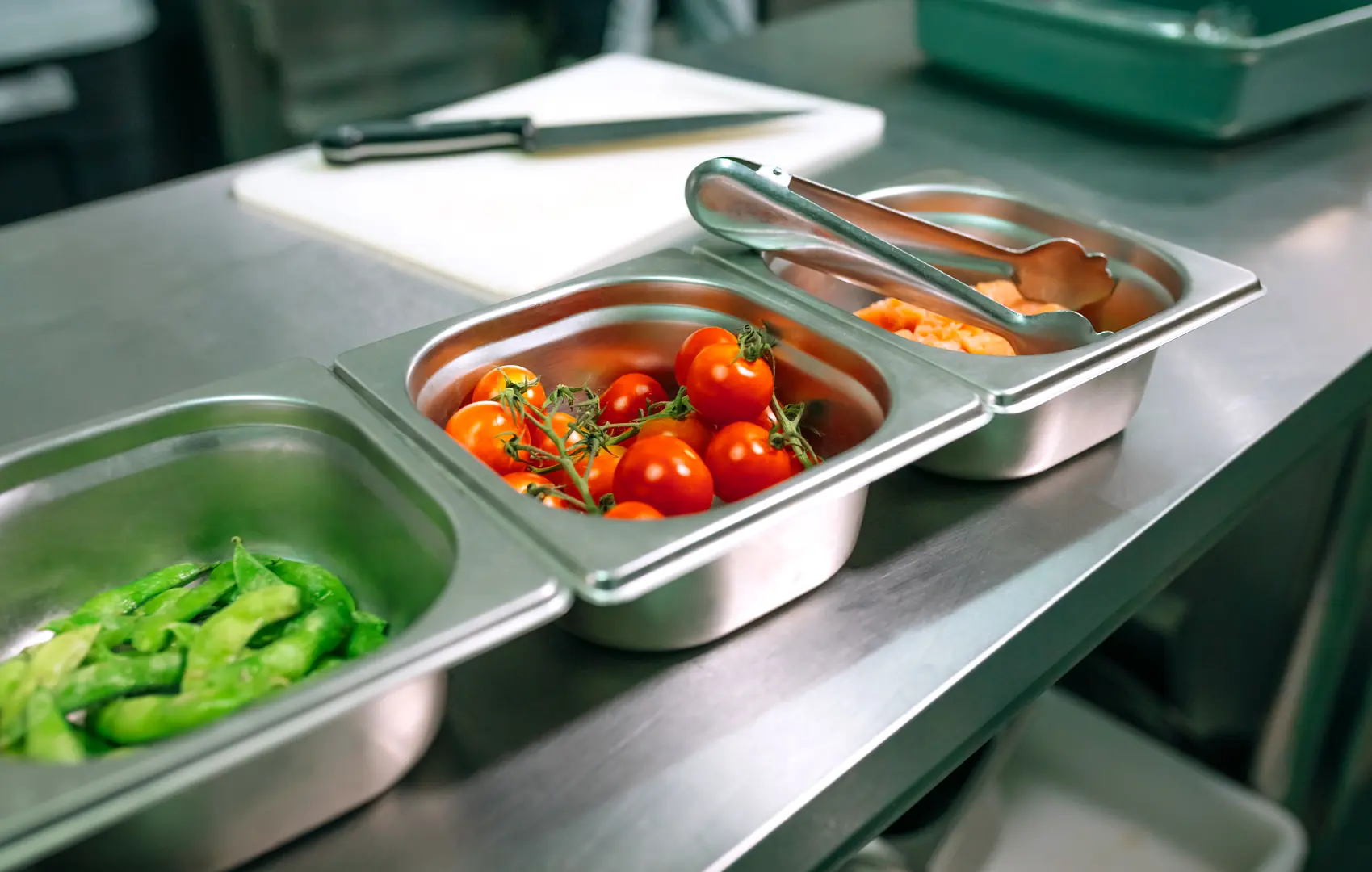 Stainless steel containers holding fresh tomatoes, sliced vegetables and kitchen utensils on a countertop.